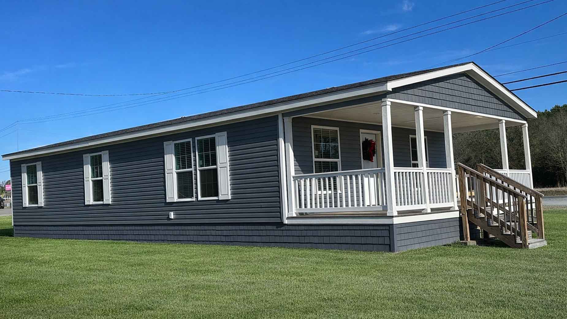 Gray manufactured home with white trim, front porch, and steps, set on a grassy lawn under a blue sky—this charming manufactured home offers comfort and style in a peaceful setting.